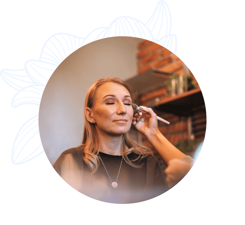 Una persona con el cabello rubio y suelto recibe un maquillaje mientras cierra los ojos, en un ambiente cálido con una pared de ladrillos al fondo.