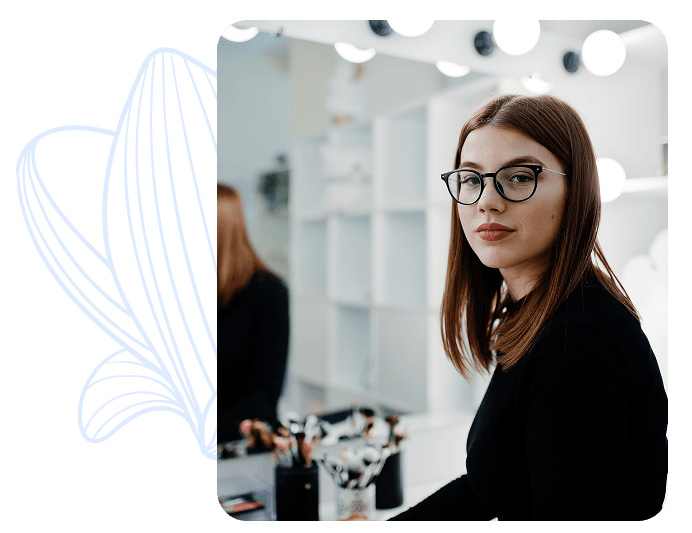 Una persona con gafas de vista posa frente a un espejo con luces de tocador, en un ambiente de maquillaje.
