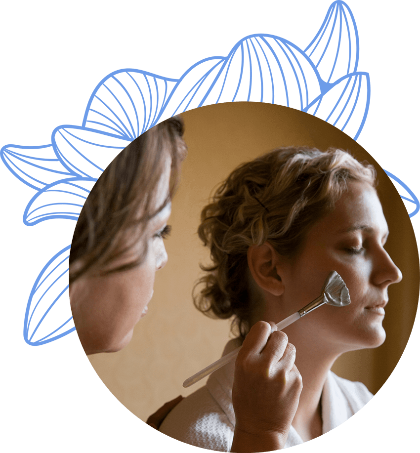 Estilista aplicando una mascarilla facial a una cliente en un entorno cálido, mientras ella se relaja con los ojos cerrados.