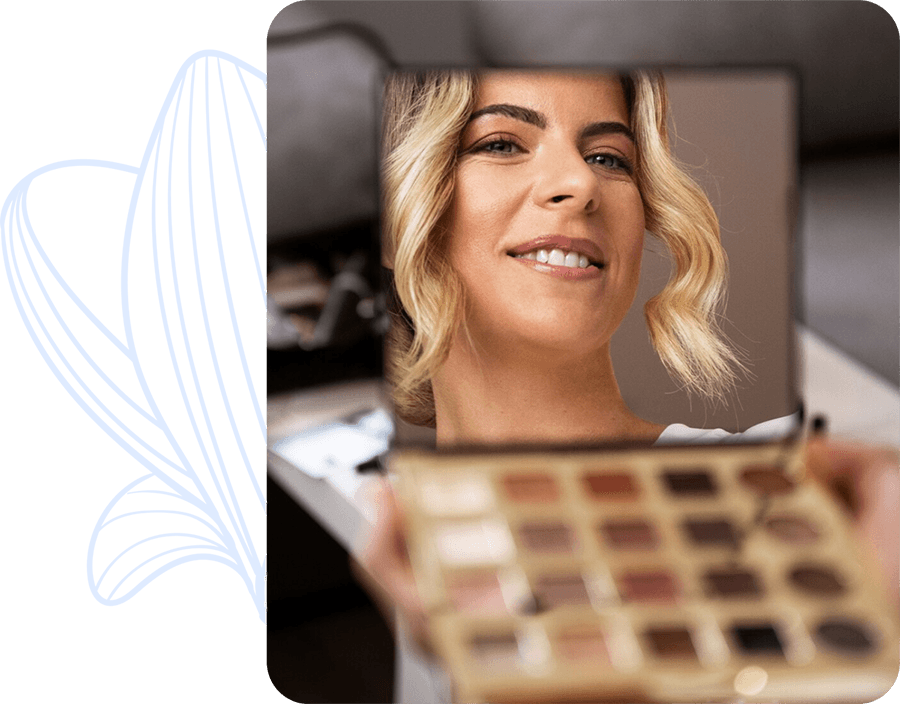 Una mujer sonríe mientras observa su reflejo en un espejo de mano, con una paleta de sombras de ojos en primer plano.