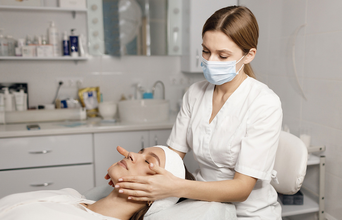 Una esteticista con mascarilla, trabajando en el rostro de una clienta que está tumbada en una camilla, en un entorno de salón de belleza con productos de cuidado facial en el fondo.