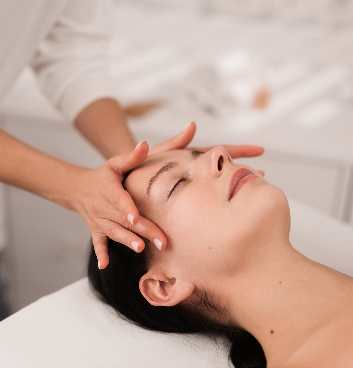 Mujer relajada disfrutando de un tratamiento facial mientras una esteticista le realiza un masaje en la cabeza en un entorno profesional.