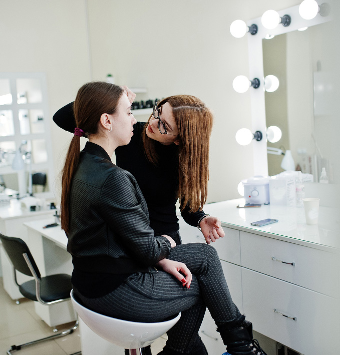 Una persona se encuentra sentada mientras otra le aplica maquillaje en un espacio iluminado por luces de tocador.