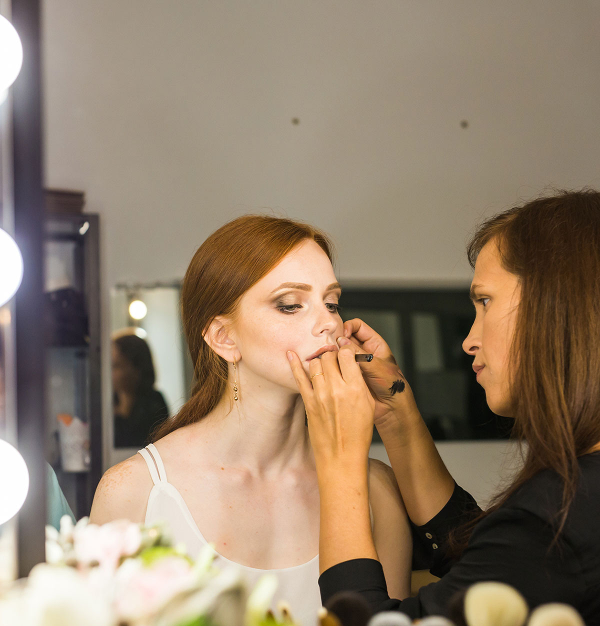 Profesional aplicando maquillaje a una modelo en un espejo de tocador bien iluminado, mientras la modelo se concentra en el proceso.