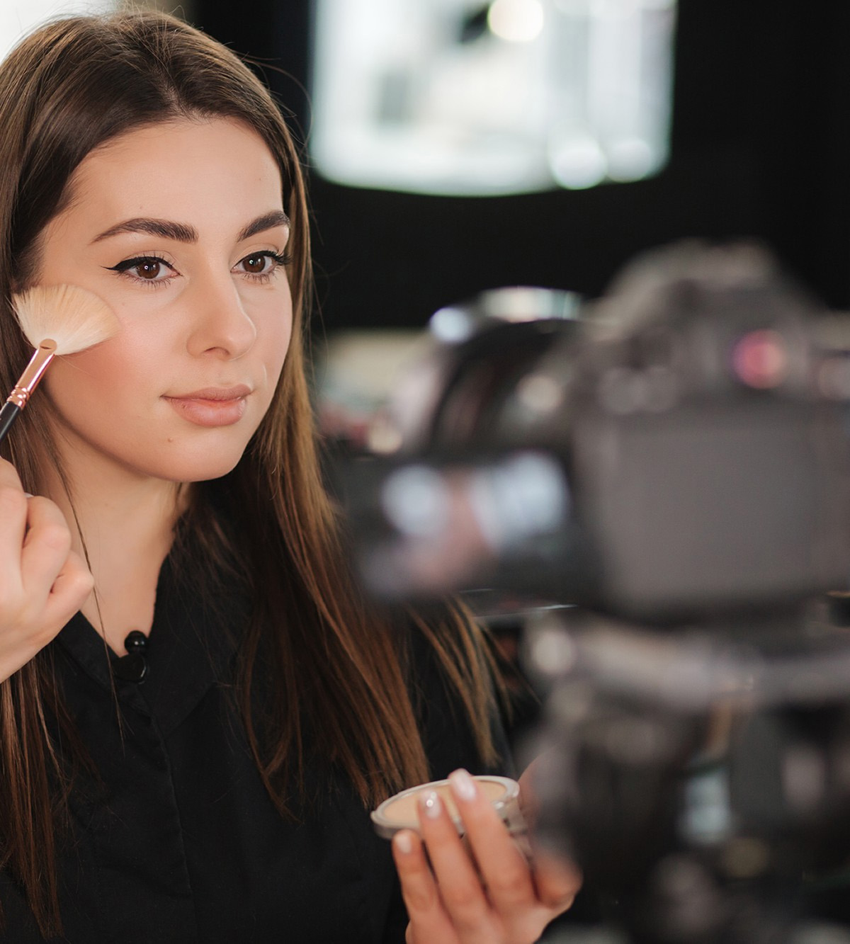 Una mujer aplica polvo facial en su mejilla mientras es grabada por una cámara, sosteniendo una caja de maquillaje en la mano.